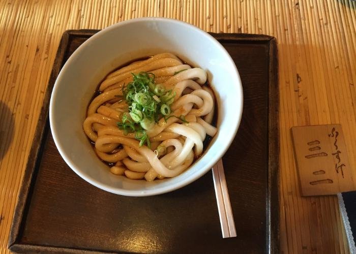 Bowl of thick udon noodles with green onion on top on brown tray with wooden chopsticks