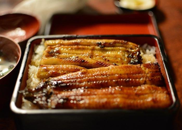 Glazed pieces of eel on rice with brown sauce in square red and black bowl