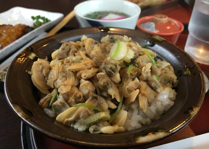 Unshelled clam meat on rice topped with green onions in brown bowl with soup in background