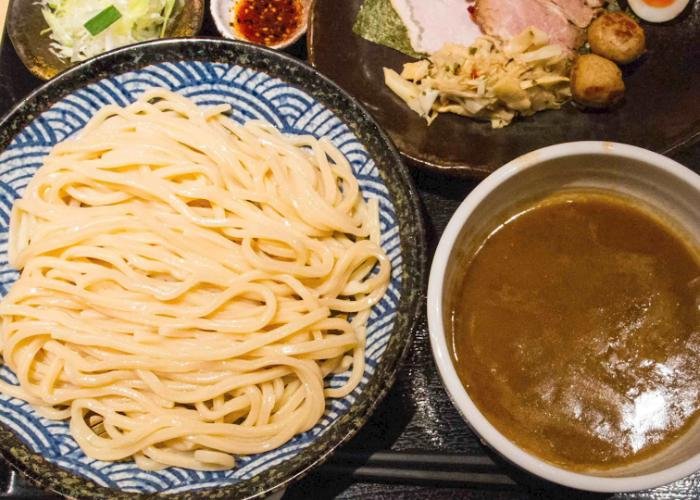 Blue dish of plain tsukemen noodles next to bowl of brown broth with other sides in background