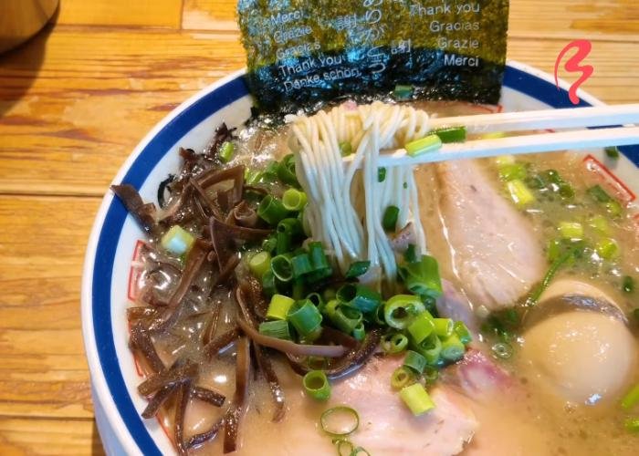 Chopsticks picking up tonkotsu ramen noodles in cloudy broth with green onions, egg, and seaweed with "thank you" written on it in different languages