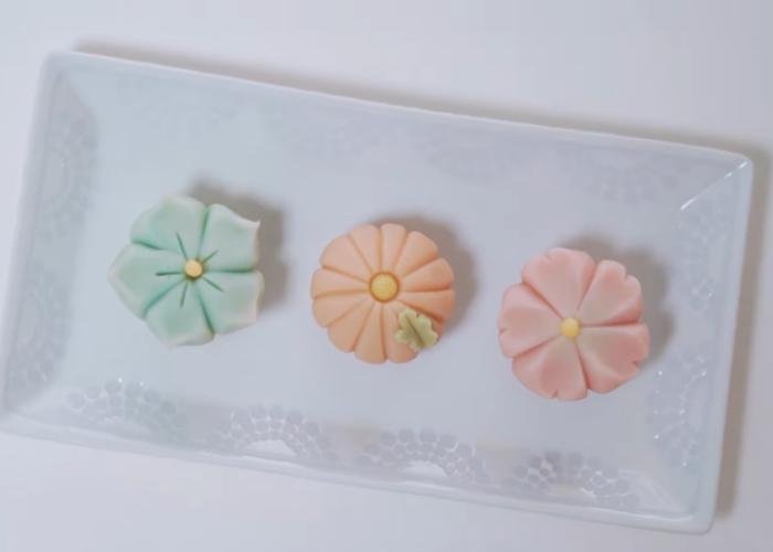 Three Japanese sweets on white plate: pink cherry blossom, orange flower, blue flower