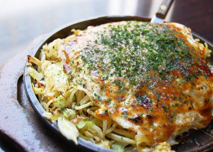A big plate of Hiroshima okonomiyaki with noodles and seafood, topped by sauce and aonori