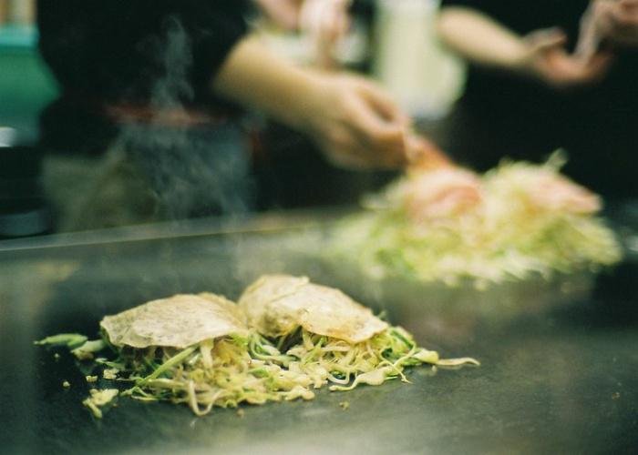 A smoking hot Hiroshima okonomiyaki, topped with vegetables, noodles and seafood, being cooked on an iron griddle