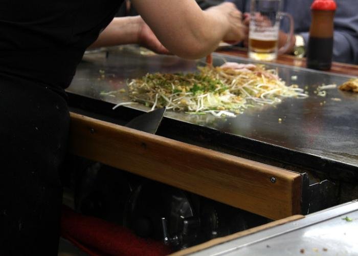 A chef adding pork to the okonomiyaki, now filled with cabbage and tenkatsu