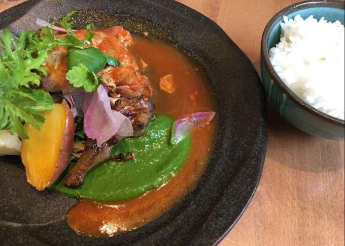 Dark brown-orange soup with vegetables and green herbs, bowl of white rice to the side