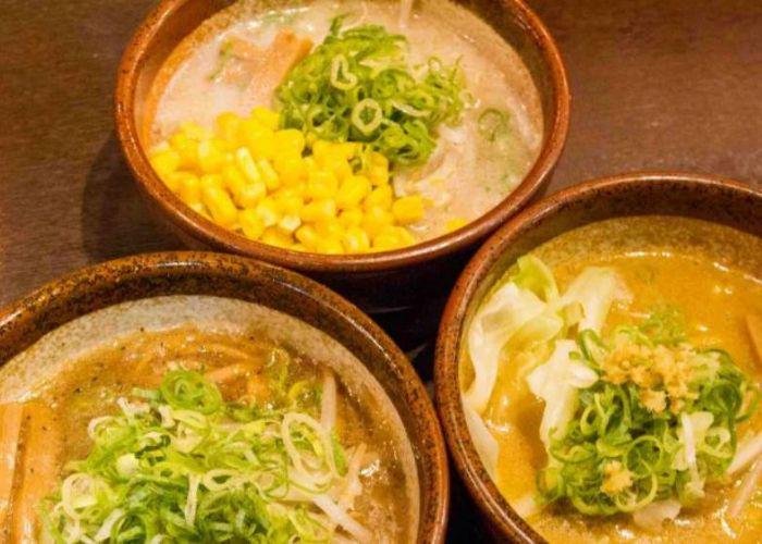 Three bowls of ramen with orange broth, topped with green onion, corn, and shredded garnishes