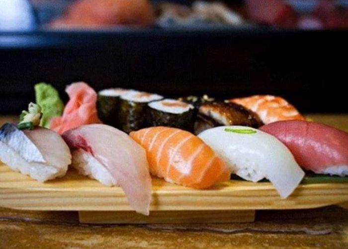 Five pieces of nigiri sushi of different bright colors on wooden platter with seaweed-wrapped rolls in background