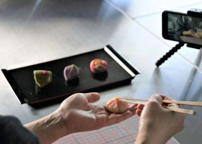 A tray of three wagashi sweets in the background, with a person's hands making another sweet and a smartphone on a stand filming it