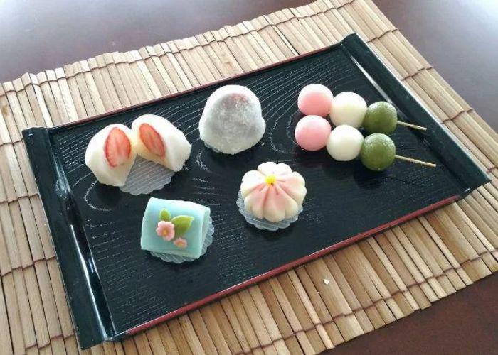 A tray of wagashi, including colorful dango, a flower-shaped sweet and ichigo daifuku, on a bamboo mat