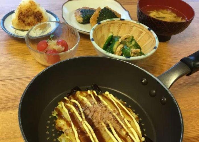 Okonomiyaki in a frying pan in the front of the shot, with several other side dishes in the background including miso soup, a rice ball and salad