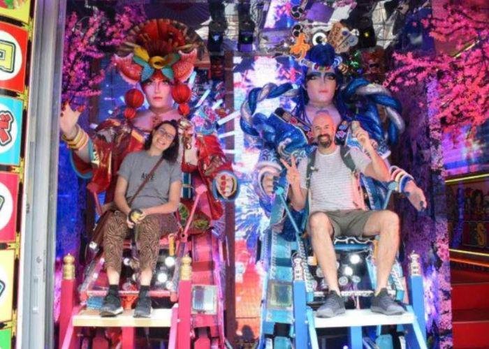 Two people in colorful neon seats with animatronic figures behind them with red and blue headpieces