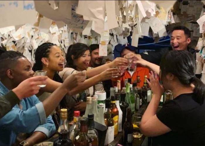 6 people and a bartender clinking glasses of beer in narrow bar with bottles of alcohol on counter