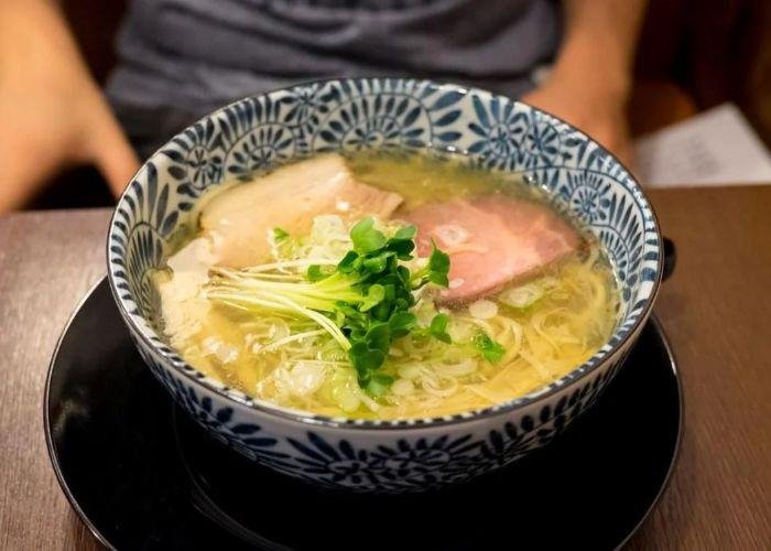White and blue patterned ramen in bowl with light broth, green garnish, and meat inside, with person's hands in background