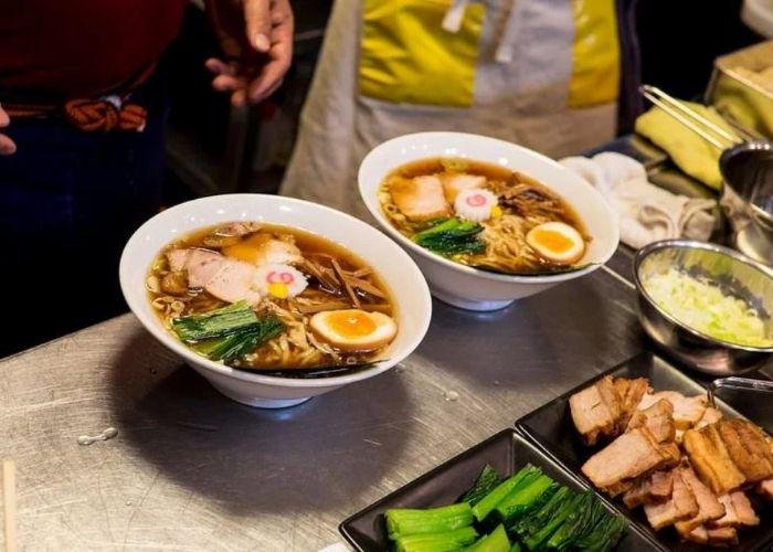 Two white bowls of dark broth ramen topped with naruto, egg, greens, meat; dishes of toppings on counter