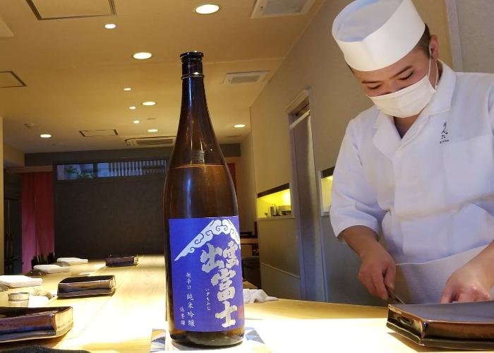 Sake bottle on the counter with a sushi chef preparing the next bite behind it