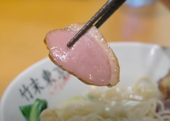 A close up image of a pair of chopsticks holding a slice of duck meat, with a bowl of ramen in the background