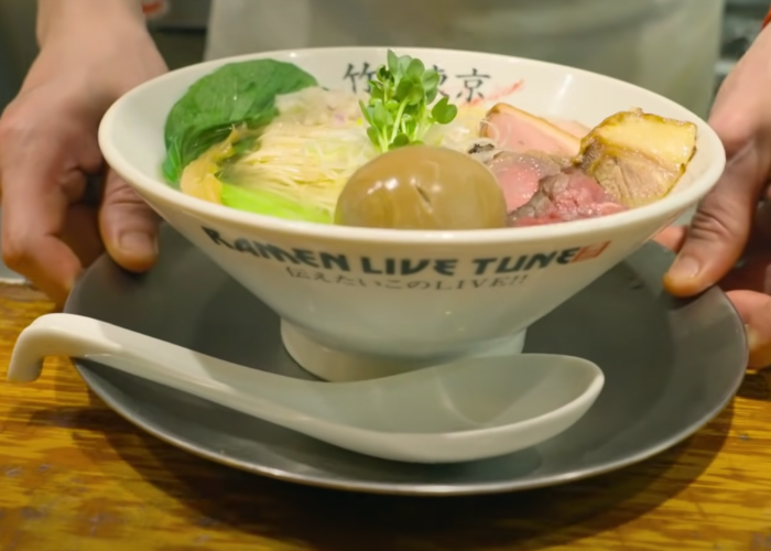 Two hands presenting a bowl of ramen on a dish with a spoon