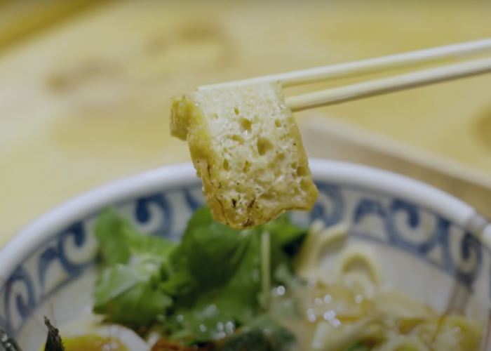 Close up shot of a pair of chopsticks holding a piece of deep-fried tofu above a bowl of ramen