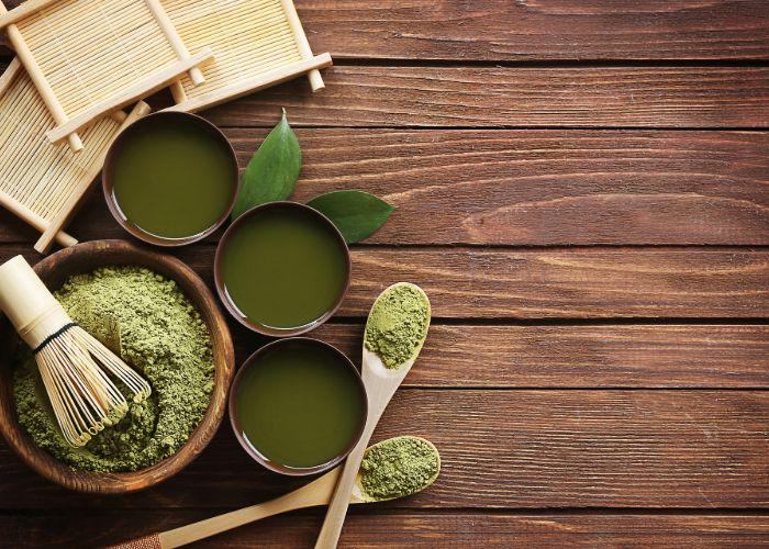 An overhead image of a bowl of green matcha powder with a whisk on top, and three cups of matcha tea