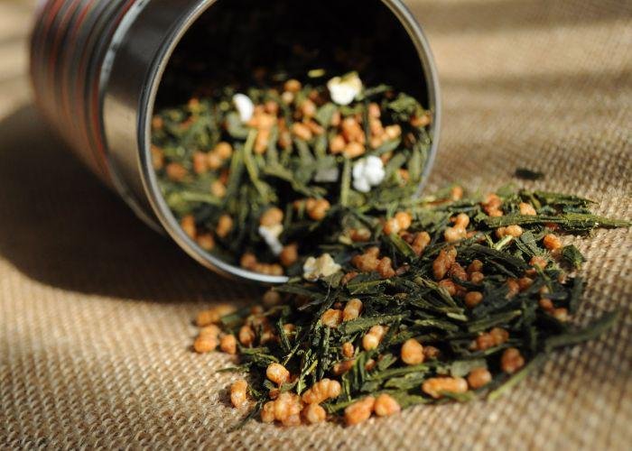 A close up image of a tin of genmaicha tea spilling out onto cloth, with dried tea leaves and roasted rice