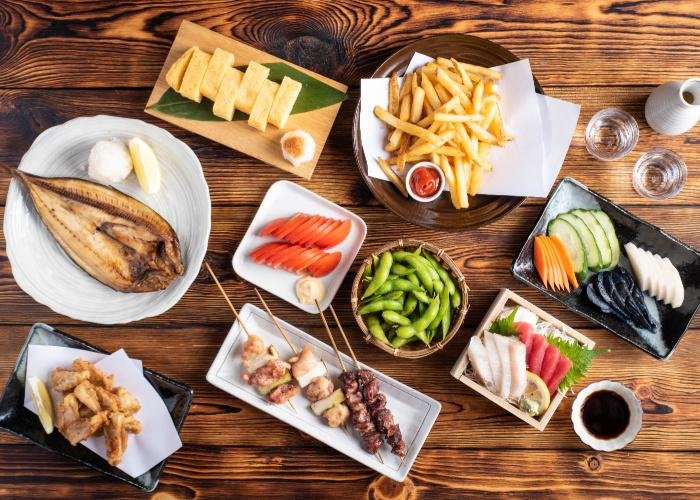 A spread of izakaya appetizers including tamagoyaki, chicken wings, grilled fish, kamaboko, sashimi and more