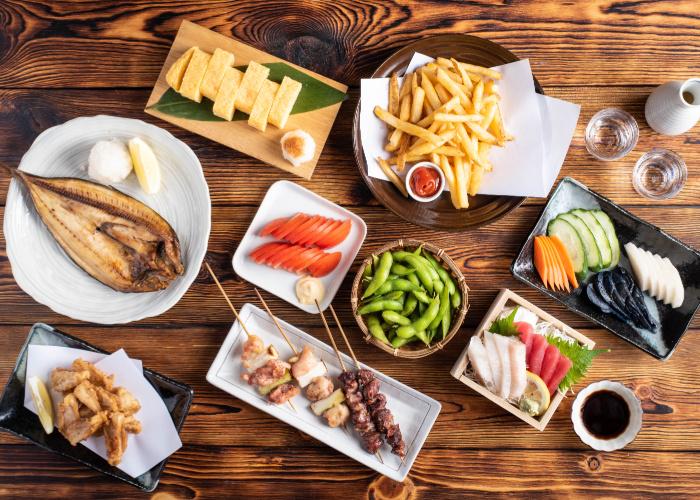 A spread of izakaya appetizers including tamagoyaki, chicken wings, grilled fish, kamaboko, sashimi and more