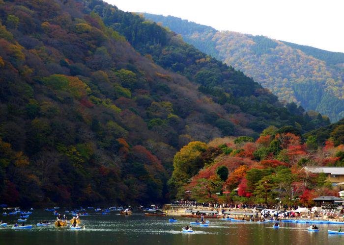 People riding boats on the river in Arashiyama with the forested mountains behind covered in red and orange trees