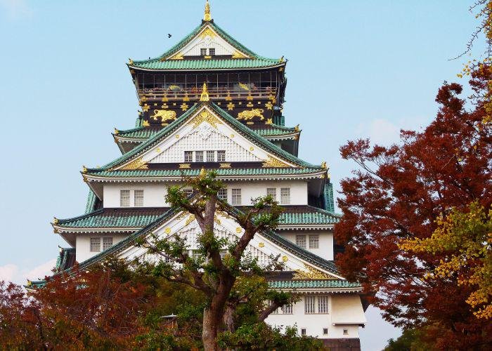 The green, white and black tower of Osaka Castle with red and gold treetops in the foreground