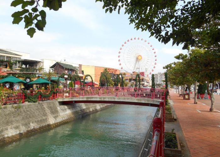 Daytime photo of American Village with the Ferris wheel in the background and a bridge over the river to shops and bars