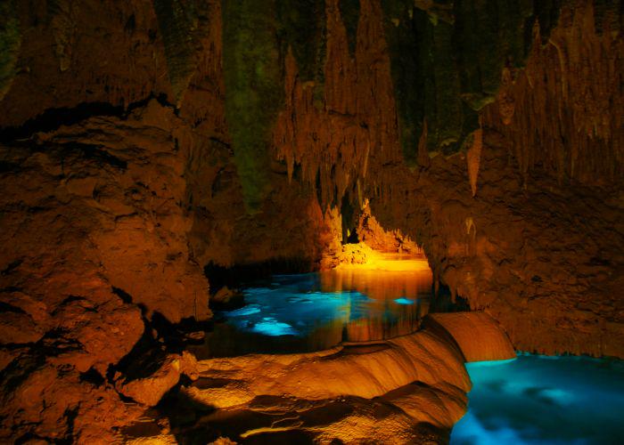 The interior of a limestone cave lined with stalactites and stalagmites and blue water at the bottom