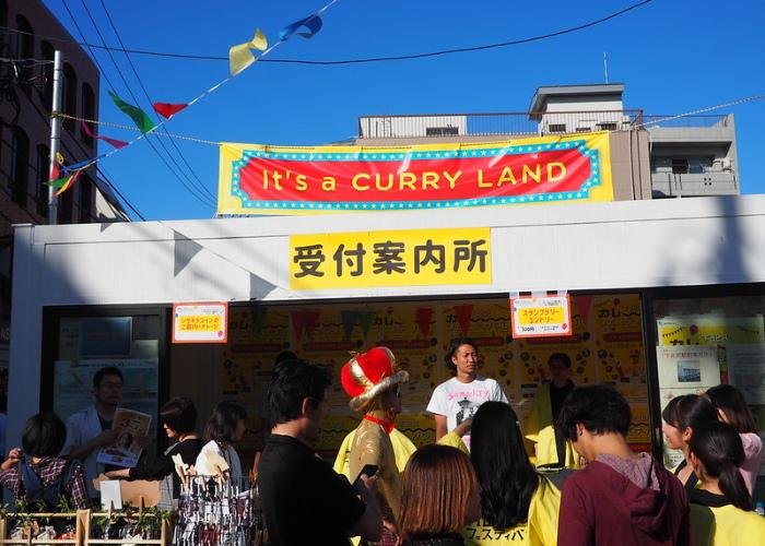 People line up outside of a curry stand at the Shimokitazawa Curry Festival, a sign reads "It's a curry land"