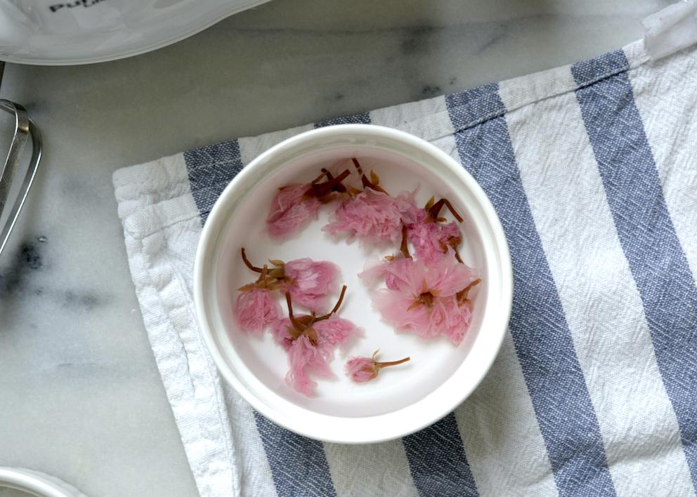 Bowl of pickled sakura blossoms floating in water on the backdrop of a striped tea towel