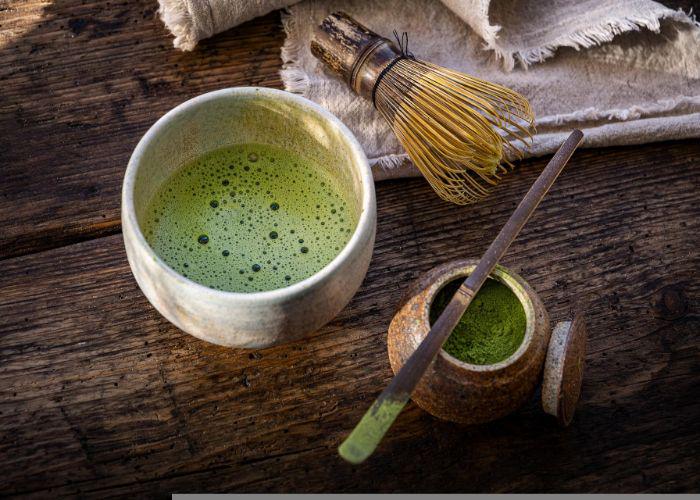 A frothy bowl of green matcha tea on a wooden table, next to a bamboo chasen whisk and a pot of bright green matcha tea powder