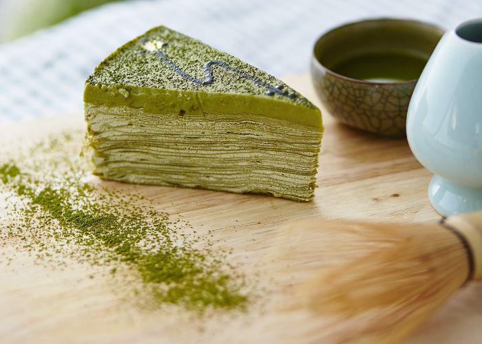 A slice of matcha cake with powdered matcha around it and a chasen whisk in the foreground