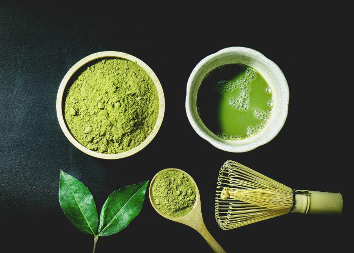 An overhead image of a bowl of green matcha powder and a bowl of matcha tea, with tea leaves and a bamboo whisk