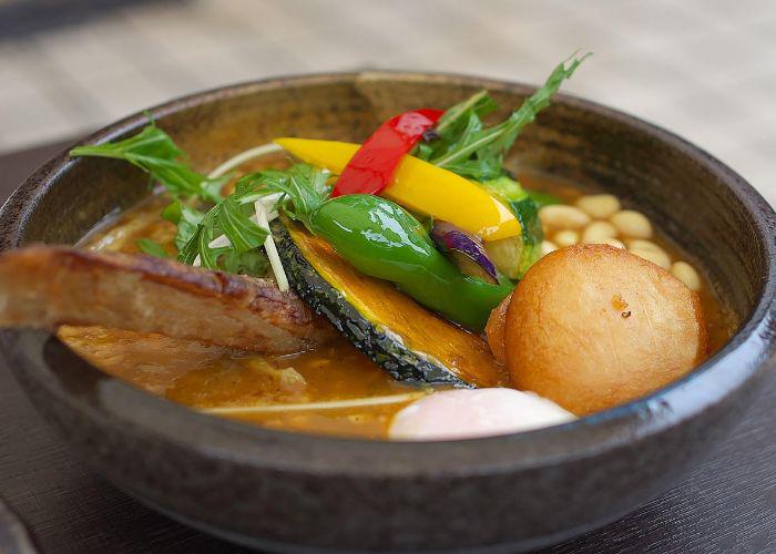 Close-up of a bowl of soup curry with vegetables including pumpkin, potato, peppers, salad leaves, beans and meat.