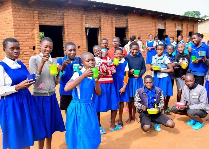 a large group of primary school students are smiling and eating their school meals before classes begin