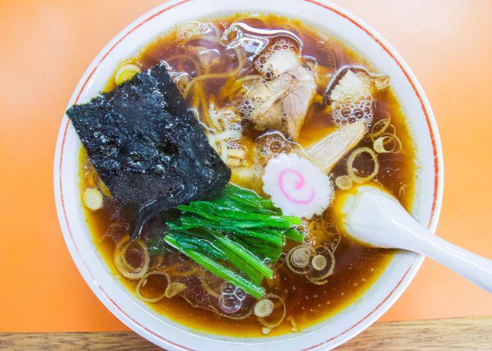 An overhead image of a bowl of ramen with a dark brown broth, big piece of seaweed, green leaves, white fishcake with a pink swirl and noodles