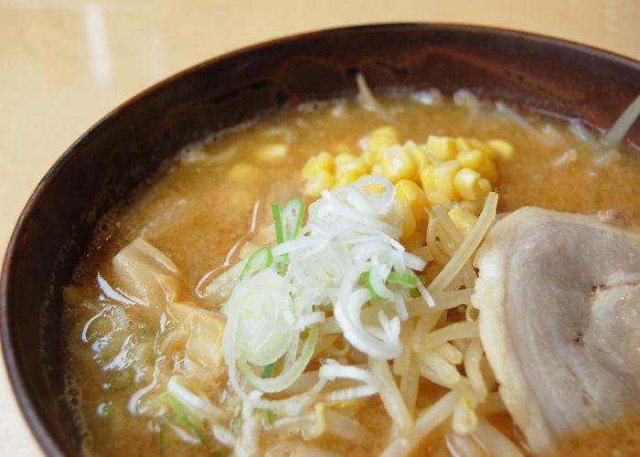 A close up image of three-quarters of a bowl of noodles, with a creamy brown broth, green onions, sweetcorn and a slice of meat