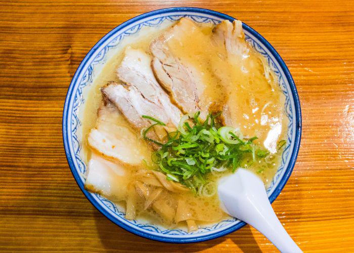 Overhead image of a bowl of noodles in an oily light brown broth, with a pile of green spring onion slices and several slices of meat