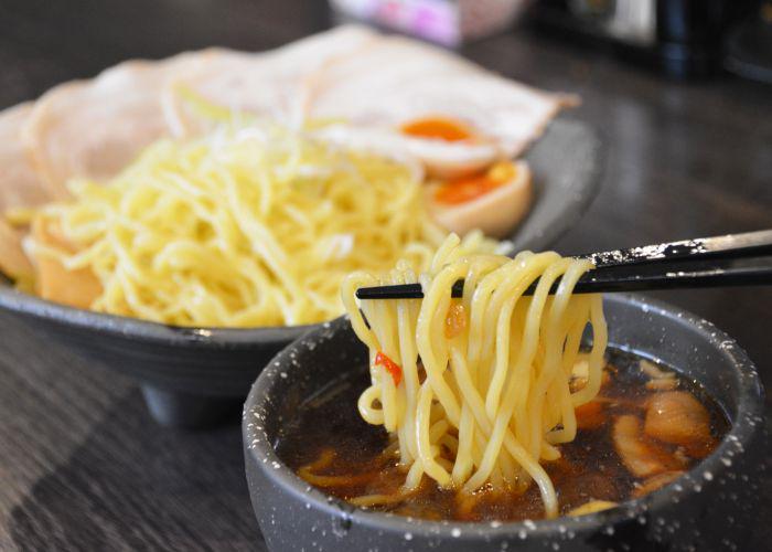 Chopsticks holding a portion of noodles and dipping them into a bowl of dark brown broth, with a larger bowl of plain noodles in the background