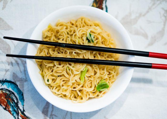 Overhead image of a bowl of curly golden instant ramen noodles with some green leaves, and a pair of black and red chopsticks resting on top