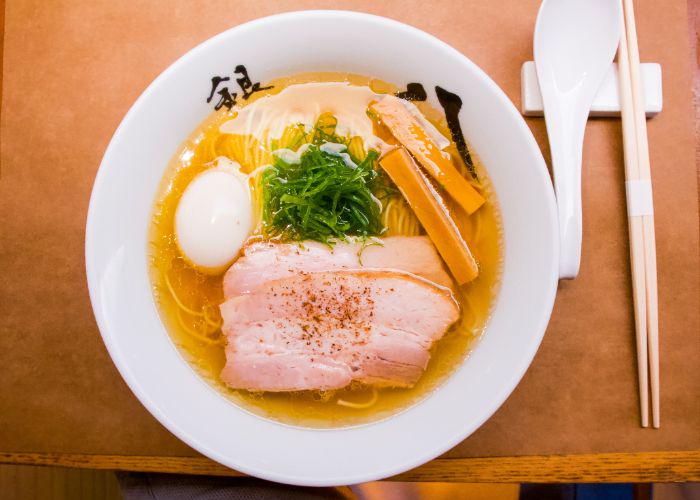 An overhead image of a white bowl of ramen noodles, with a clear golden broth, whole white egg, slices of meat, and a pile of green seaweed
