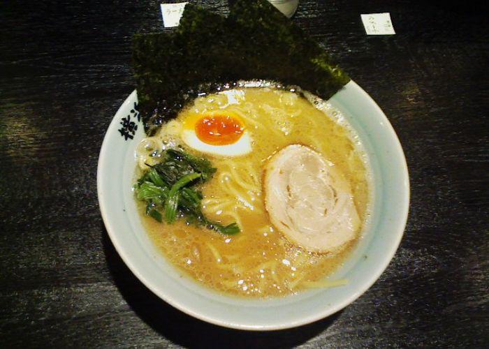 An overhead shot of a bowl of ramen with an oily golden broth, topped with half an egg, a slice of meat, greens, and sheets of seaweed lining one side of the bowl