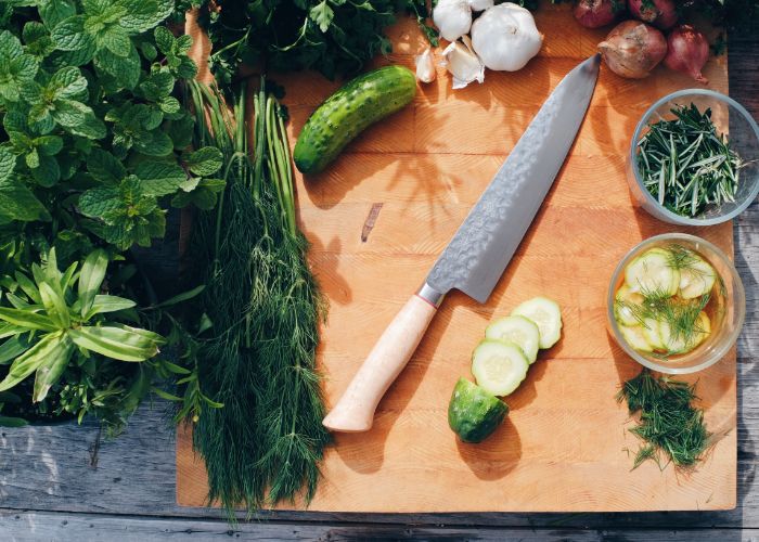 Overhead image of a gyuto knife on a wooden chopping board surrounded by slices of cucumber and herbs