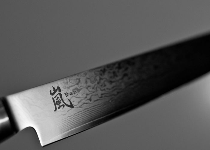Close up image of the blade of a Japanese knife against a grey background, with a kanji printed on the side