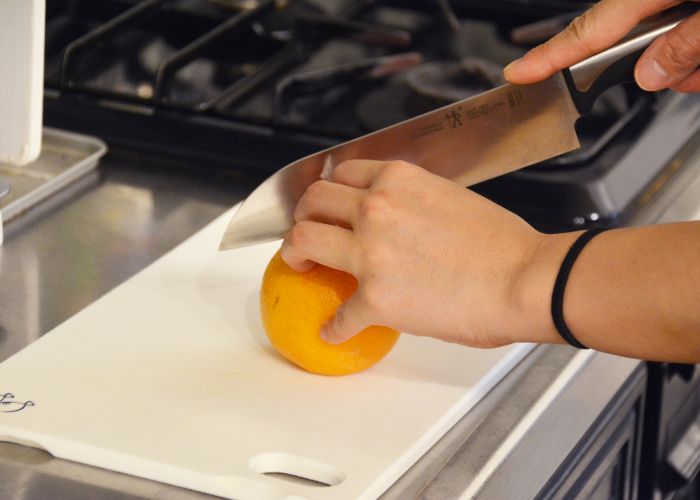 A close up image of a person's hands holding a knife and cutting into an orange on a white plastic chopping board