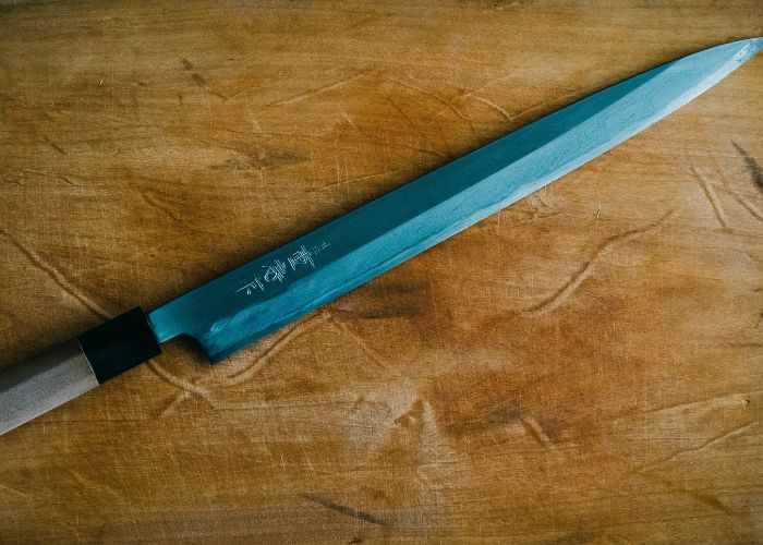 An overhead image of a long, thin knife resting on a wooden table, with Japanese kanji on the blade