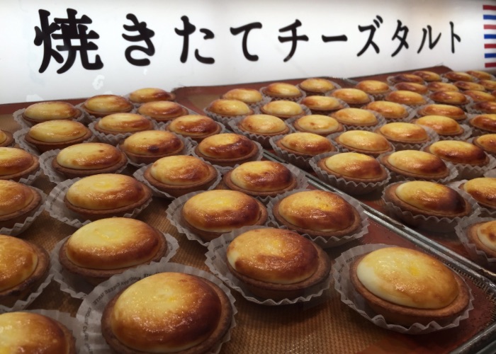 Freshly-baked cheese tarts lined up at a Kinotoya bakery. An illuminated sign behind reads in Japanese, "Freshly-Baked Cheese Tarts".